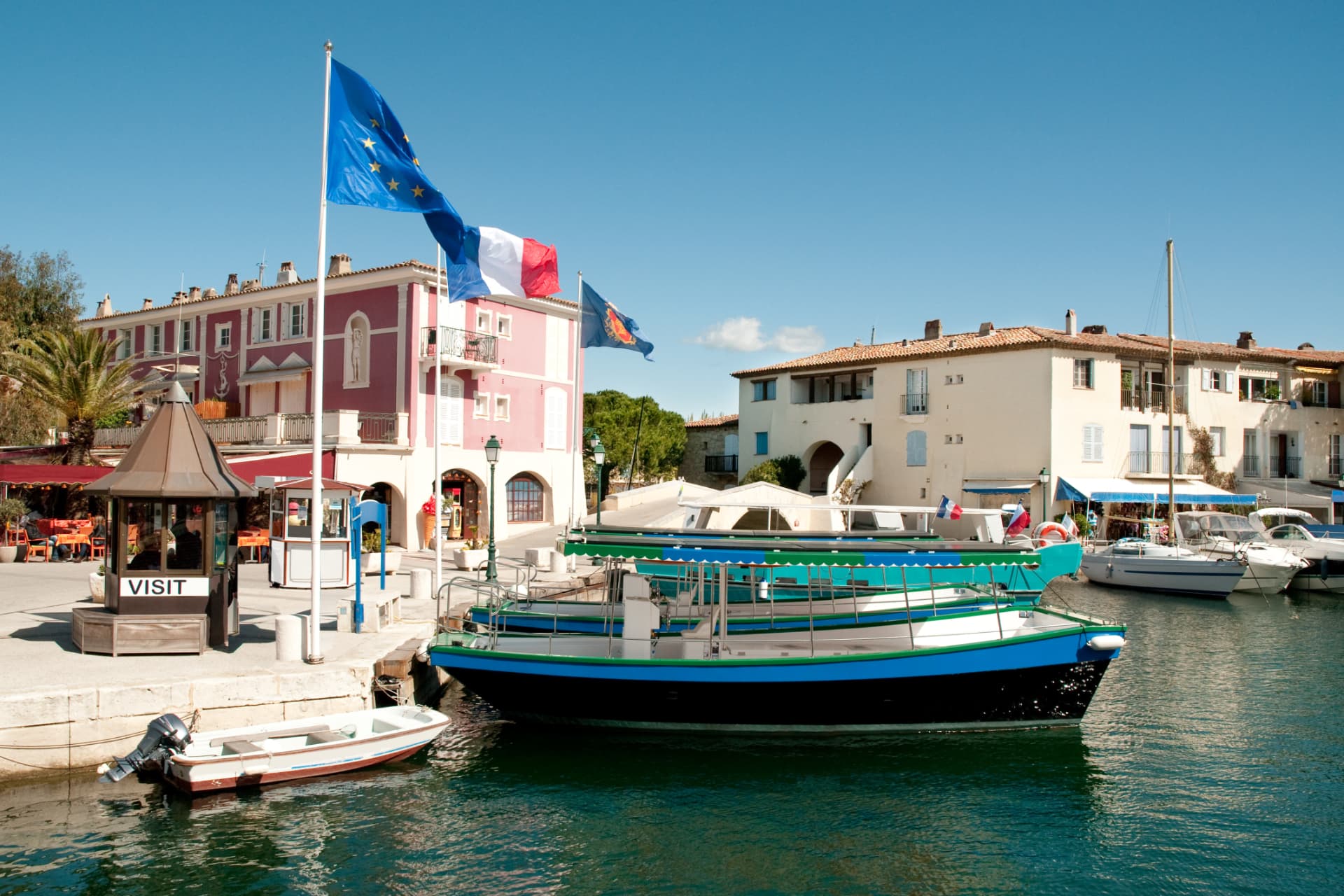 Quai de Port Grimaud avec drapeaux européens et français, maisons colorées et bateaux amarrés